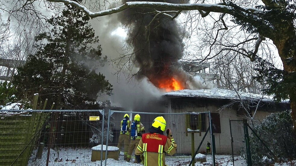 Außenansicht eines eingeschossigen Hauses, aus dessen schneebedecktem Dach Flammen aufsteigen. Im Vordergrund Einsatzkräfte der Feuerwehr, drum herum winterliche Bäume.