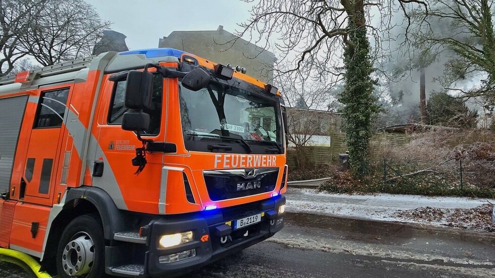 Ein Feuerwehrauto steht an einer Straße. Im Hintergrund steigt über Gebäuden Rauch auf.