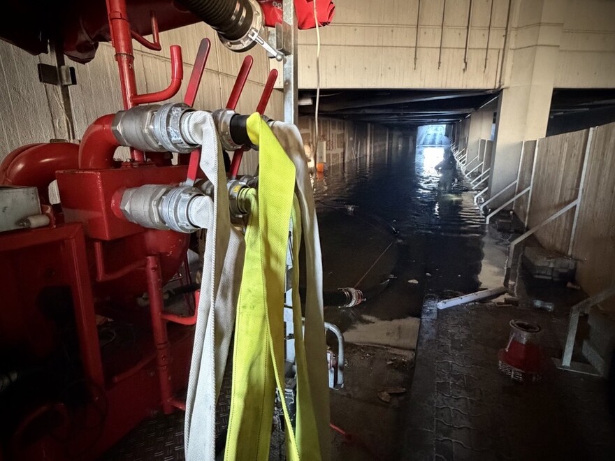 Blick in weitläufige Kellerräume, deren Boden mit Wasser überlaufen ist. Im Vordergrund eie Aufhängung für Schläuche.