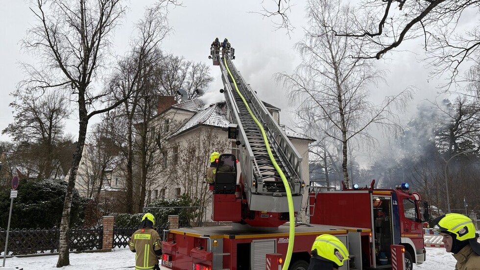 Iim Vordergrund ein Feuerwehrauto mit ausgeschobener Drehleiter, in deren Korb zwei Retter mit einem Schlauch stehen. Sie löschen das Feuer an einem Haus im Hintergrund; Rauch zieht dort entlang. Das Haus steht in verschneiter Landschaft.