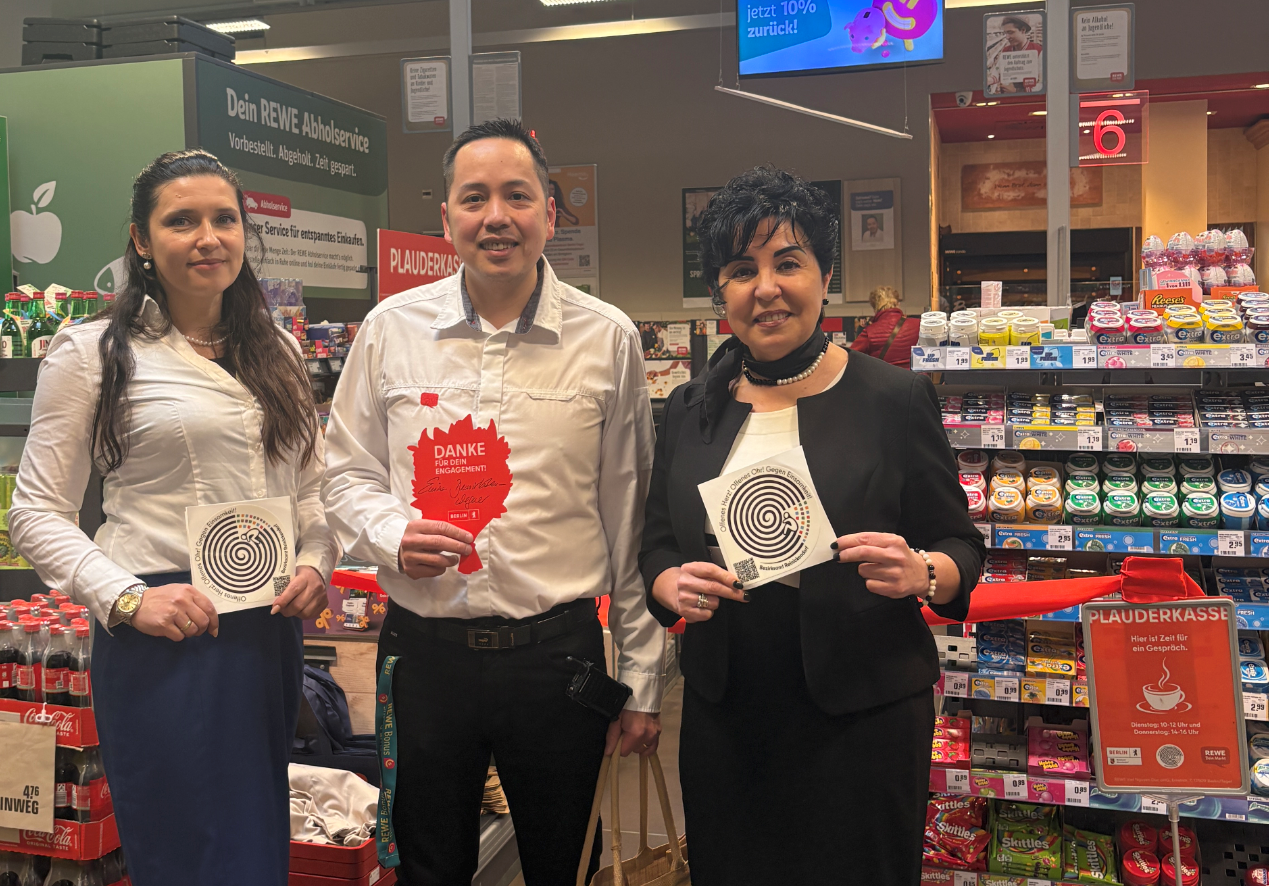 drei Menschen in einem Supermarkt, die in die Kamera schauen. Im Hintergrund der Kassenbereich.