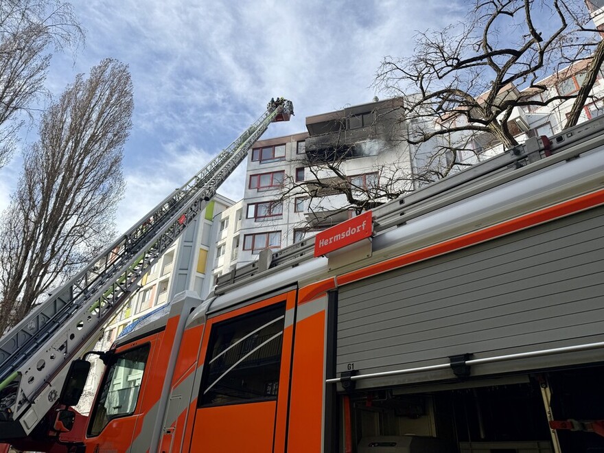 Im Vordergrund ein Feuerwehrauto mit ausgefahrener Drehleiter. Diese lehnt an einer Hauswand eines Mehrfamilienhauses. Aus den Fenstern eines oberen Stockwerks dringt Rauch.