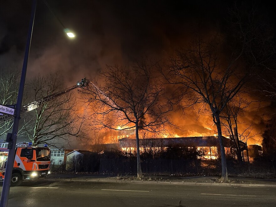 Das nächtliche Foto zeigt Feuerschein und Rauch über einem großen Gebäude. Im Vordergrund eine baumgesäumte Straße