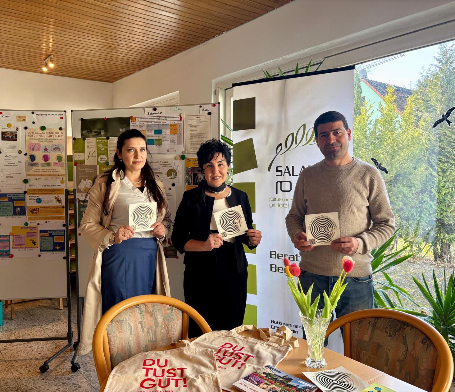 Drei Menschen in einem Innenraum, halten ein Blatt Papier mit einem Symbol in die Kamera. Vor ihnen ein Tisch mit einer Blumenvase, hinten ein Fenster und Rollup-Banner