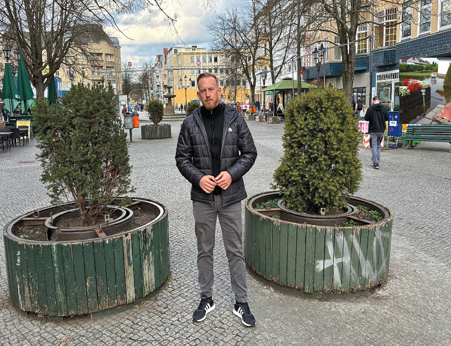 Ein Mann vor einer Bank in einer Fußgängerzone. Die Bank steht zwischen zwei Koniferen.