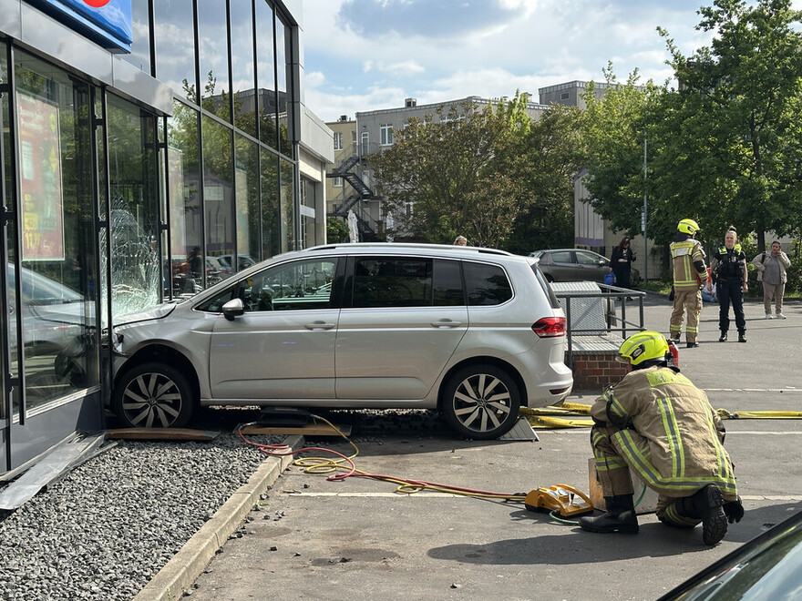 Ein Auto, das mit der Front die gläserne Fassade eines Gebäudes durchbrochen hat, davor ein Feuerwehrmann im Einsatz
