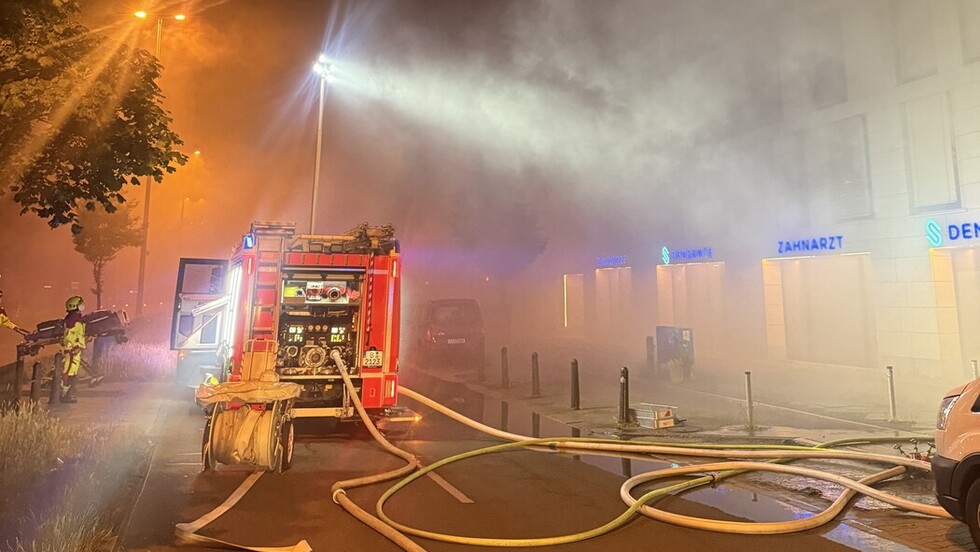 Feuerwehrfahrzeuge und Schläuche in einer Straße bei Flutlicht in der Nacht. Viel Rauch ist in der Luft.