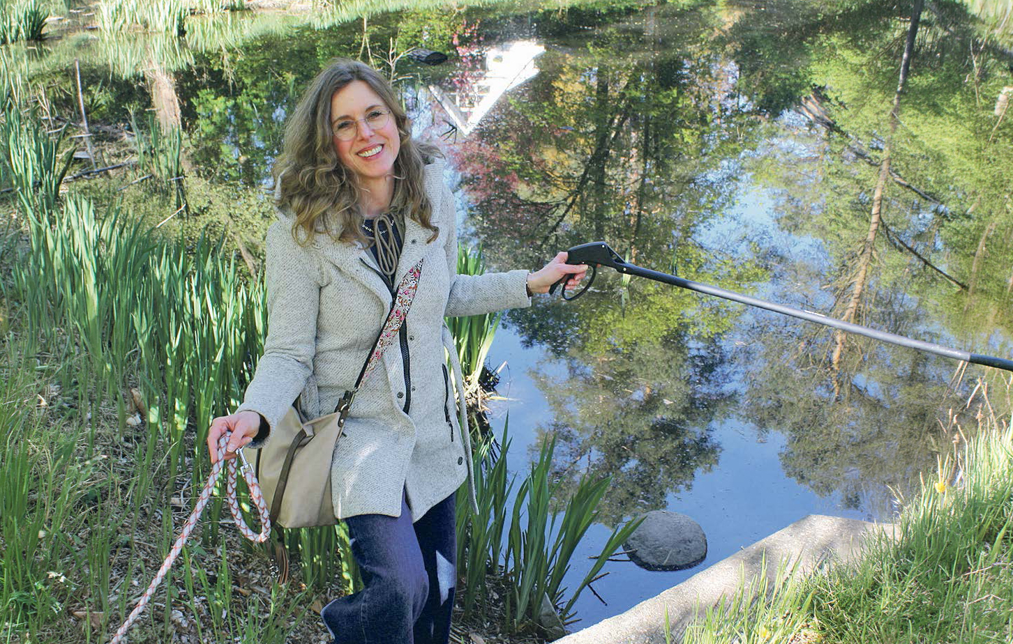 Eine Frau geht mit Leine und Greifzange an einem Teich entlang