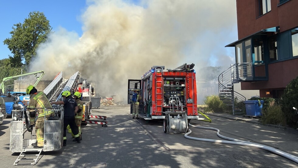 Rückansicht eines Feuerwehrautos, davor viel grauer Rauch vor blauem Himmel, einige Feuerwehrleute im Vordergrund, die an einer eingefahrenen Leiter tätig sind.