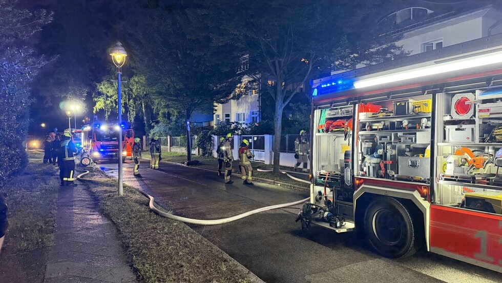 Ein Feuerwehreinsatz an einer nächtlichen Straße. Im Vordergrund ein Einsatzfahrzeug, weiter hinen ein ausgerollter Schlauch und mehrere Einsatzkräfte.