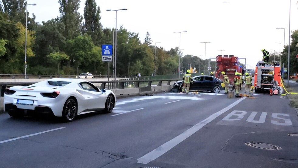 Zu sehen ist eine Unfallstelle: Feuerwehrleute arbeiten um ein verunfalltes schwarzes Auto, Bindemittel auf der Straße, im Hintergrund Einsatzfahrzeuge